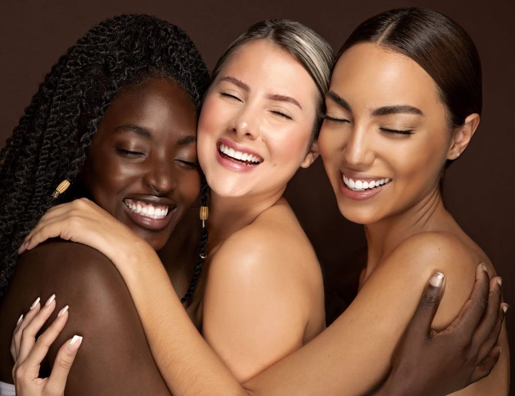 Three diverse women embracing with eyes closed and smiling, celebrating friendship and unity.