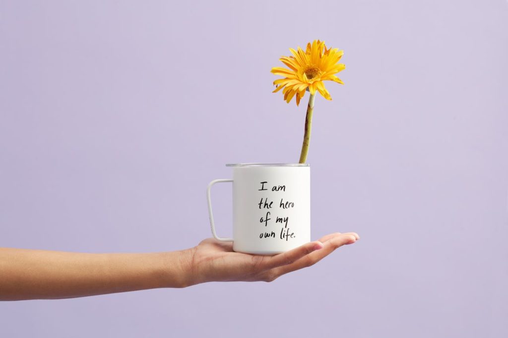 Hand holding a mug with a sunflower and motivational quote, set against a purple background.